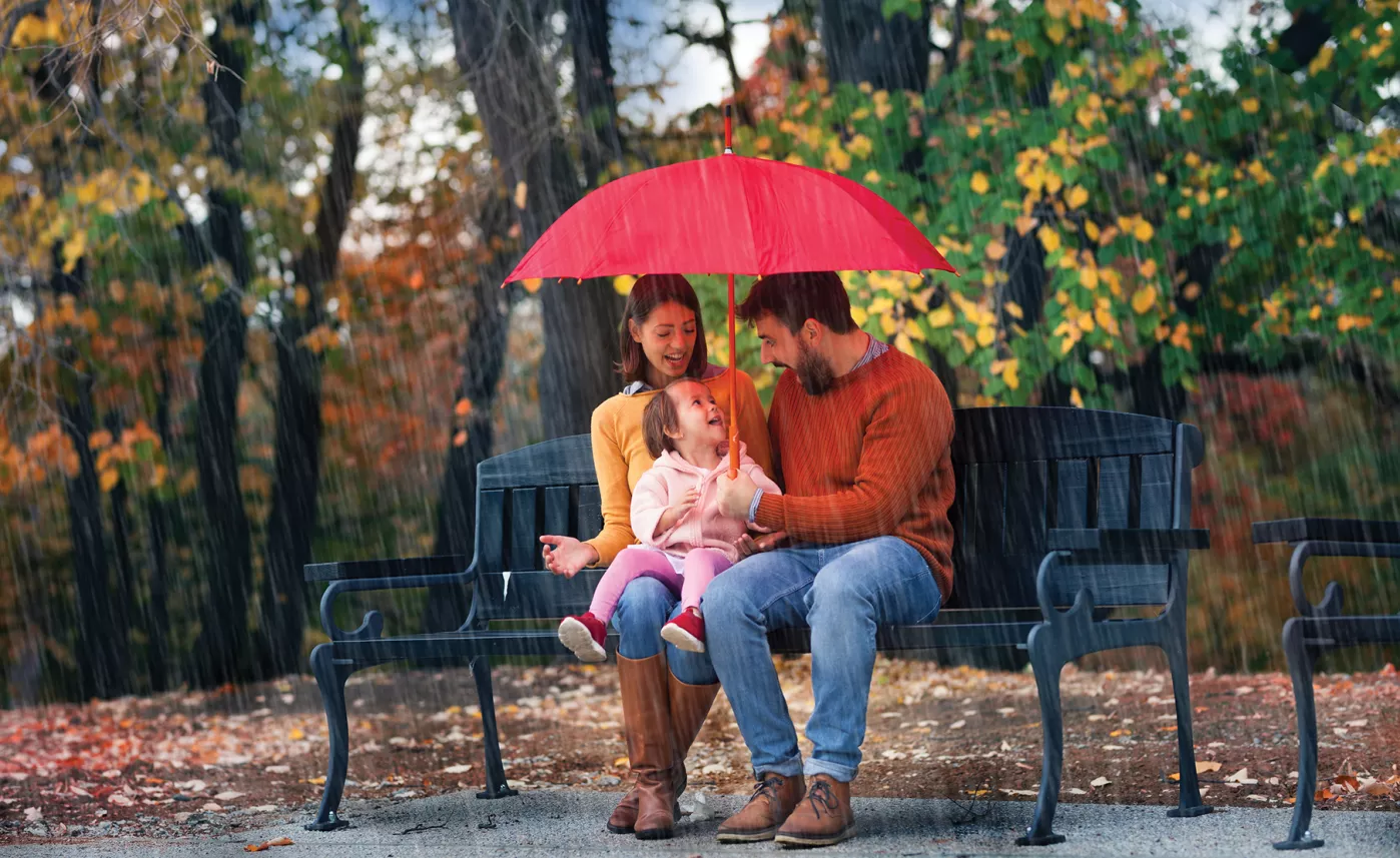  De jeunes parents s’assoient sur un banc de parc avec leur petite fille sous un parapluie par temps pluvieux.
