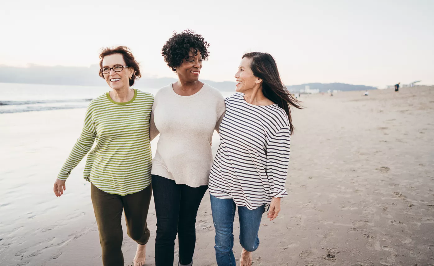  Three retirement-aged women walk together on a beach.

