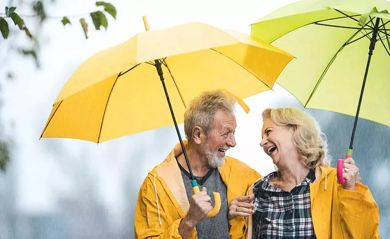  Un couple de personnes âgées rit sous la pluie, avec des parapluies et des imperméables jaunes, et profite du moment.
