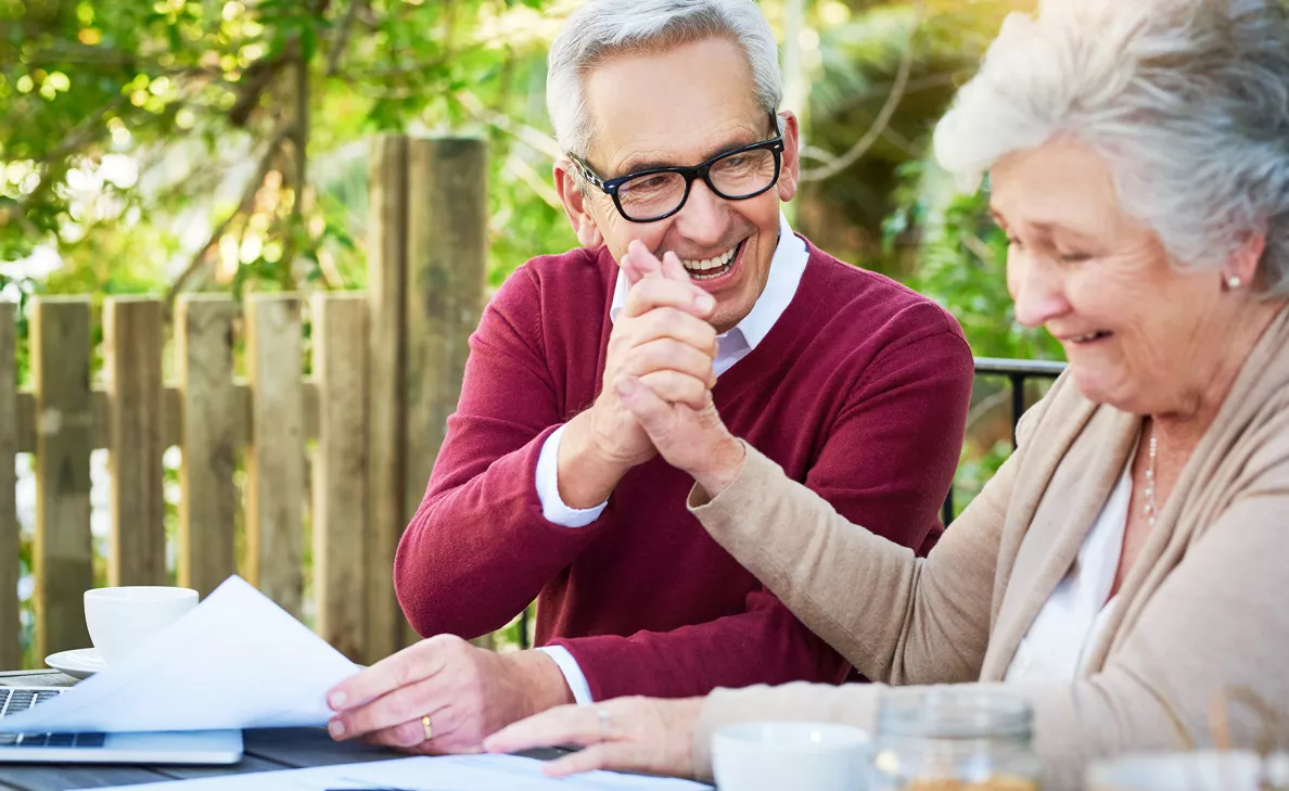  Un couple à l’âge de la retraite sourit, main dans la main, en regardant des documents sur le balcon.
