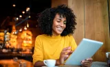Une femme sourit en lisant de l’information sur une tablette dans un café branché.